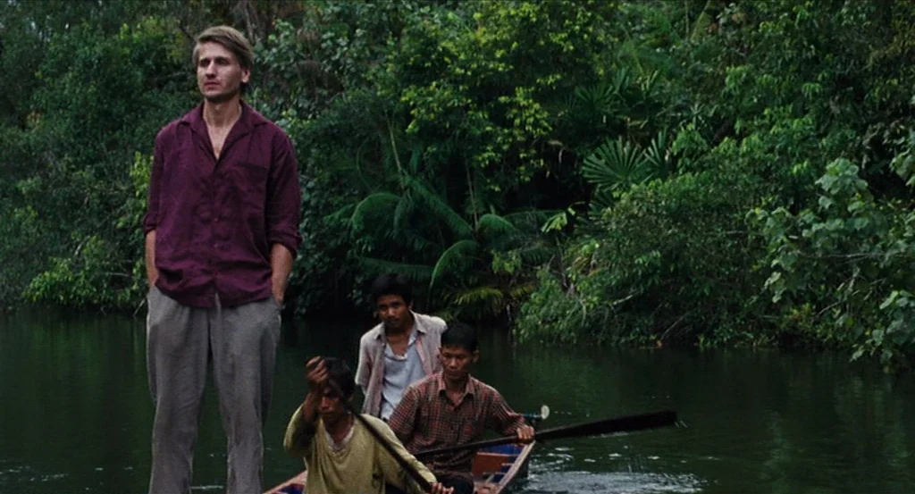Almayer’s Folly (2011) movie still: fullbody — A dense riverbank filled with lush tropical vegetation sits along a calm, dark river. One man stands…