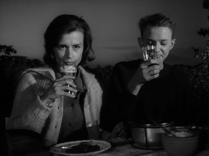 Through A Glass Darkly (1961) movie still: medium — Two people sit at a table outdoors at twilight in front of a stone wall. They both hold up…