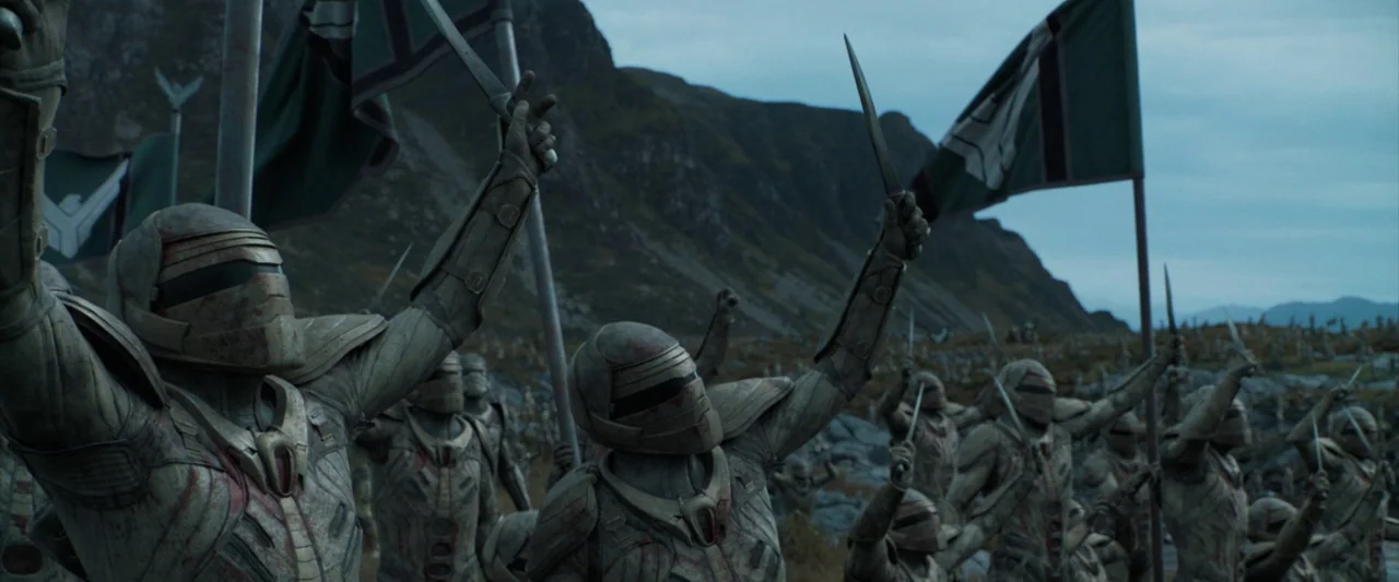 Dune (2021) movie still: medium — A group of armored soldiers stands on a craggy, rocky hillside under an overcast sky. The soldiers hold swords and…