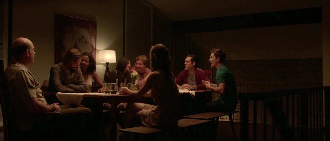 The Invitation (2015) movie still: wide — A group of friends sits at a wooden dining table in a dimly lit home. They are eating and conversing with…