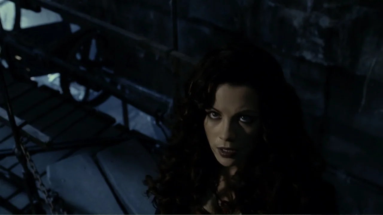 Van Helsing (2004) movie still: closeup — A dark, industrial interior space featuring metal stairs and railings. A woman with long wavy hair looks…