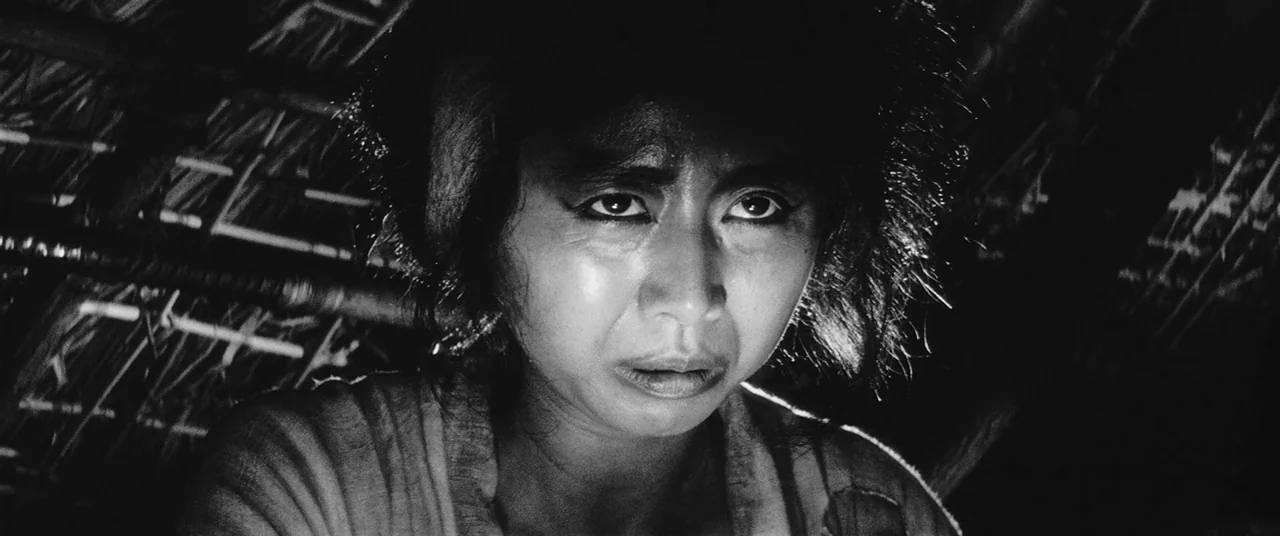 Onibaba (1964) movie still: closeup — A dark, cramped interior of a thatched hut with rough wooden beams overhead. A woman looks directly at the…