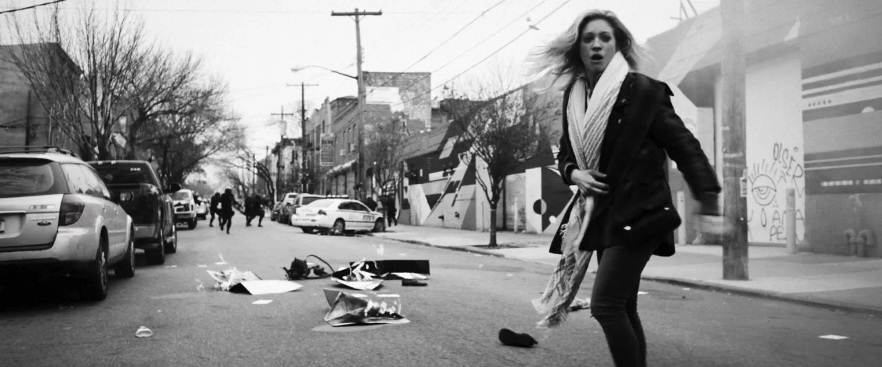 Bushwick (2017) movie still: medium — A city street with parked cars, graffiti-covered walls, and debris scattered across the asphalt. A young woman…