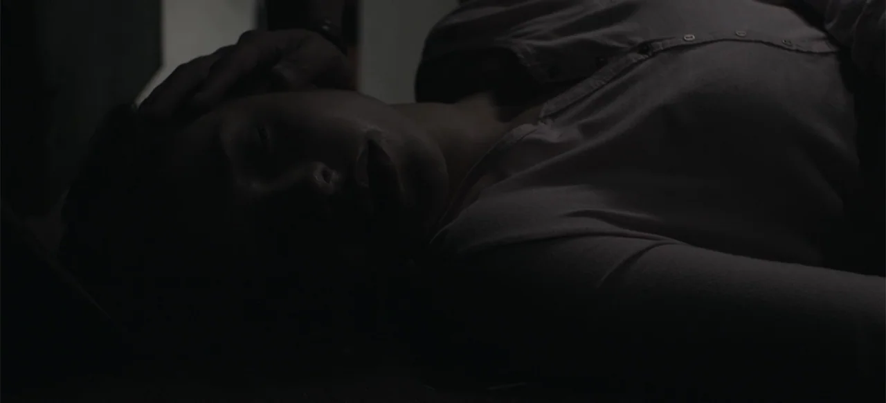 About the Deaths (2016) movie still: closeup — A dark bedroom interior shows a person lying down in low light. The subject is resting on a surface…
