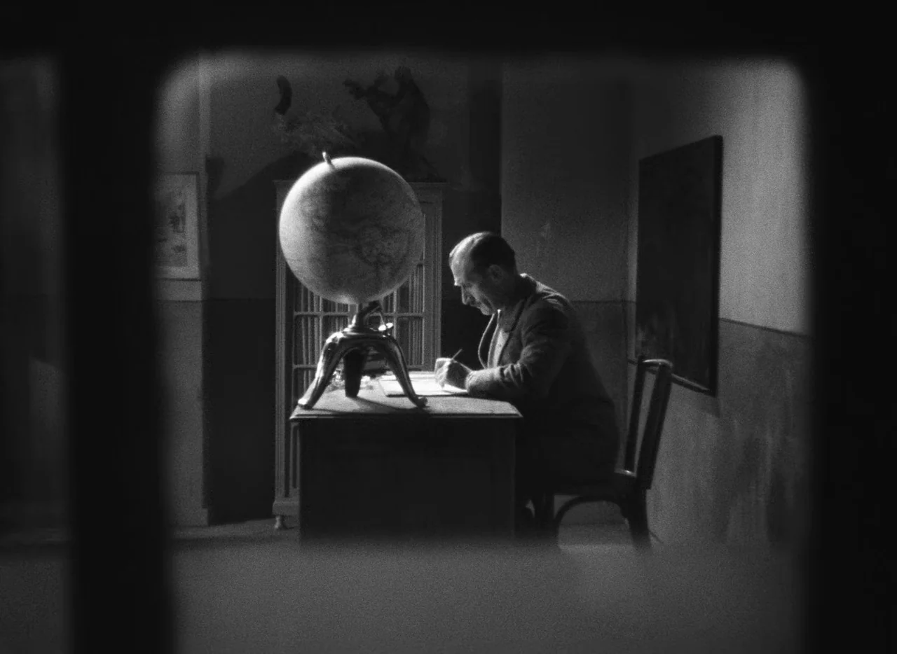 Le Corbeau (1943) movie still: medium — A dimly lit room features a man sitting at a wooden desk at night. A large globe rests on the desk next to a…