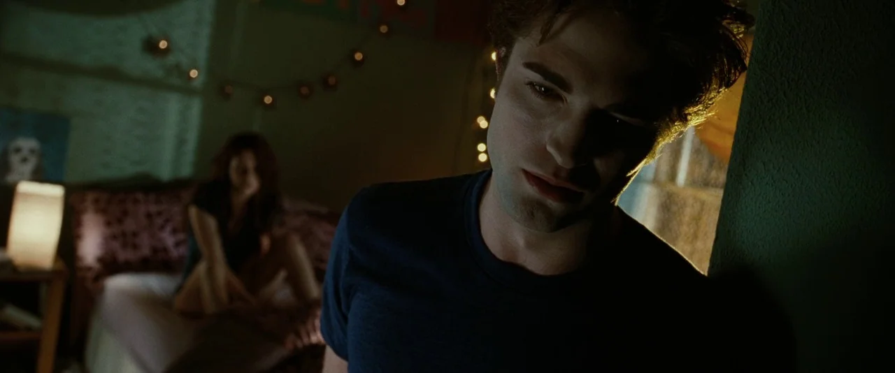 Twilight (2008) movie still: medium — A dimly lit room features a man in the foreground and a woman on a bed. String lights hang on the wall, and…