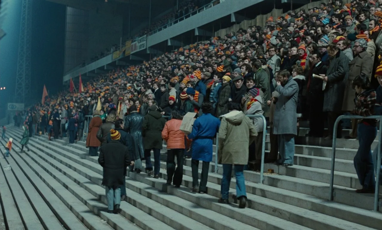 Graduate First (1978) movie still: wide — A vast, crowded concrete stadium section is filled with spectators in winter attire. Many people move up…