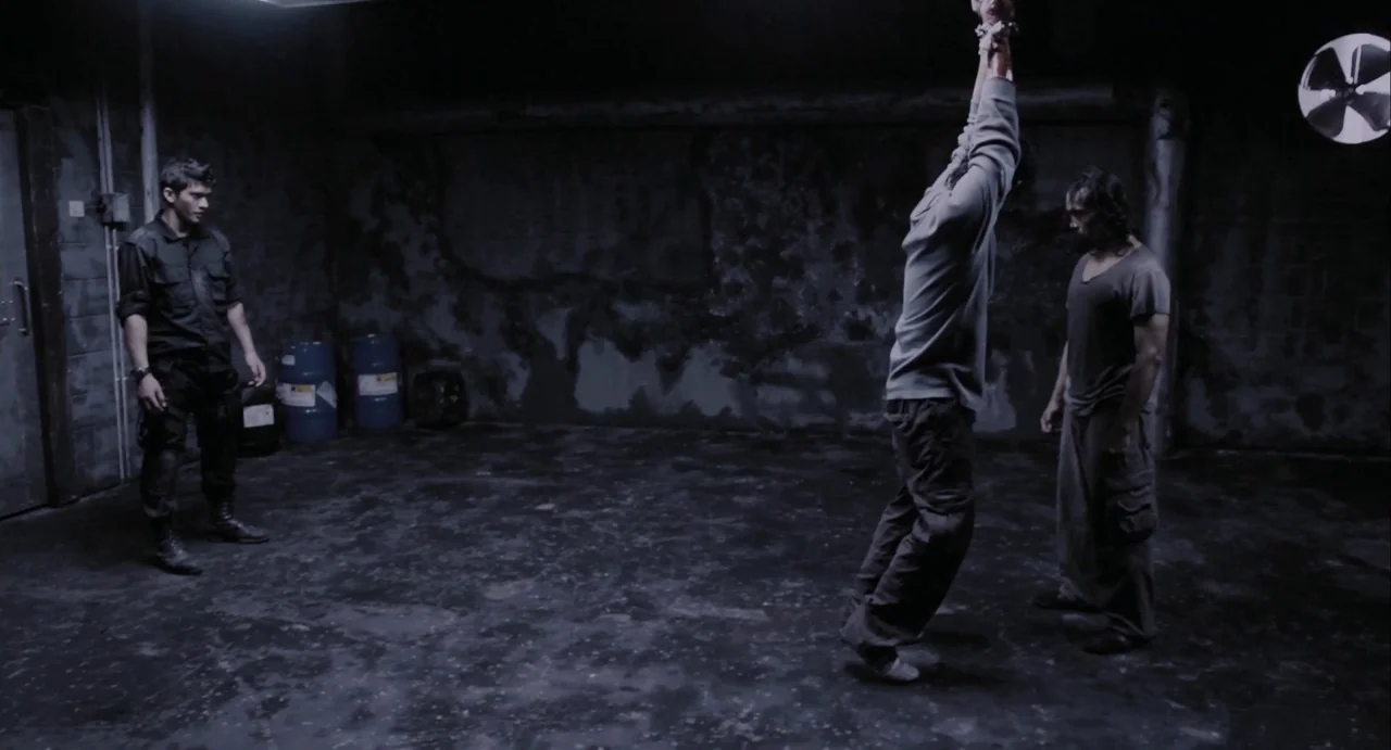 The Raid (2012) movie still: wide — A dimly lit industrial room with exposed concrete walls and two large blue barrels. One person is suspended by…