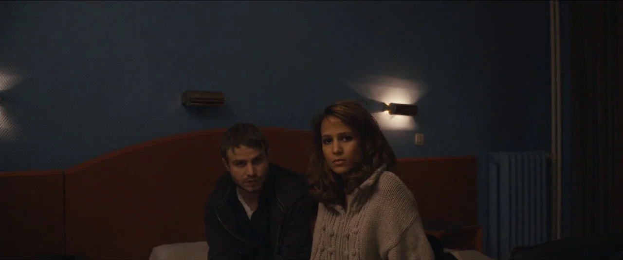 Simon Killer movie still: medium — A dimly lit hotel room features a dark blue wall and an orange headboard. A young man and a woman sit…