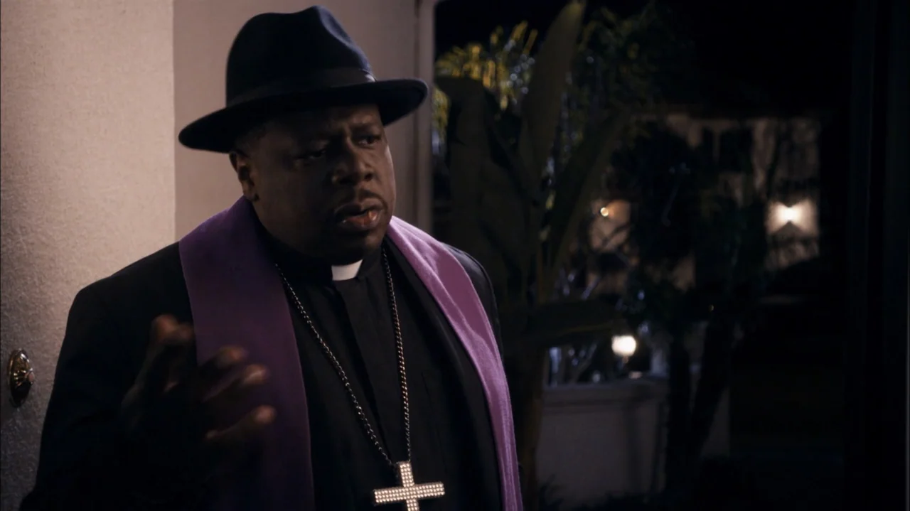 A Haunted House (2013) movie still: medium — A man wearing a clerical collar and purple stole stands on a dark porch. He is speaking while gesturing…
