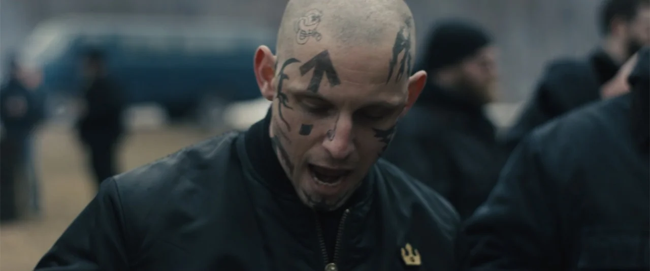 Skin (2009) movie still: closeup — An outdoor area with a blurred blue vehicle visible in the distance. A man with heavy face tattoos shouts while…