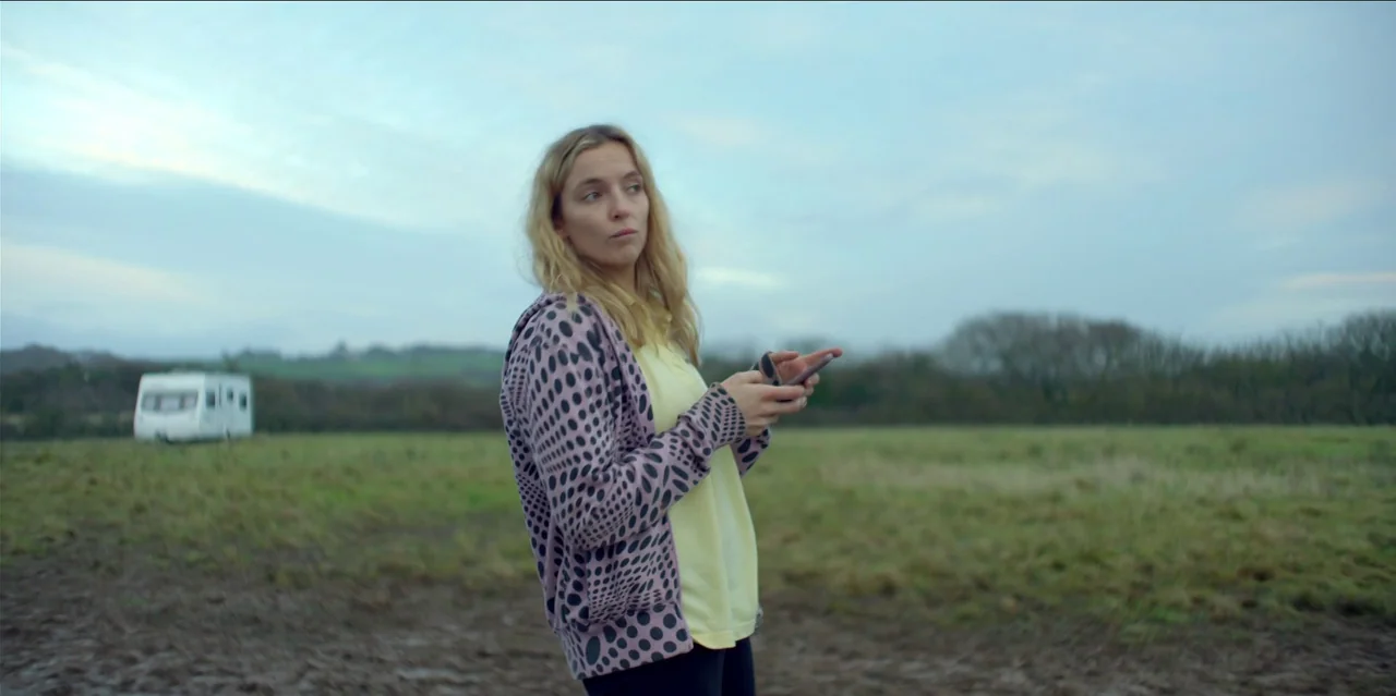 Help (2021) movie still: medium — A vast, overcast field features a distant white travel trailer under a pale sky. A young woman in a patterned…