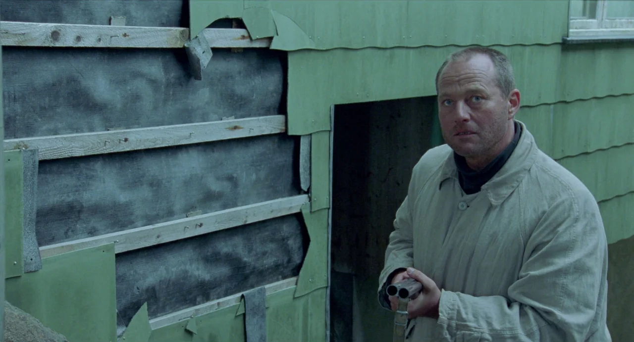 Insomnia (1997) movie still: medium — A green wooden wall with peeling paint and exposed black underlayment and wooden studs. A man holds a shotgun…