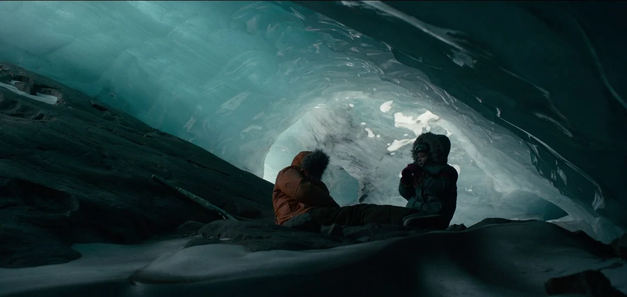 The Midnight Sky (2020) movie still: wide — A cavern formed within deep blue glacial ice with smooth, undulating surfaces. Two people in winter…