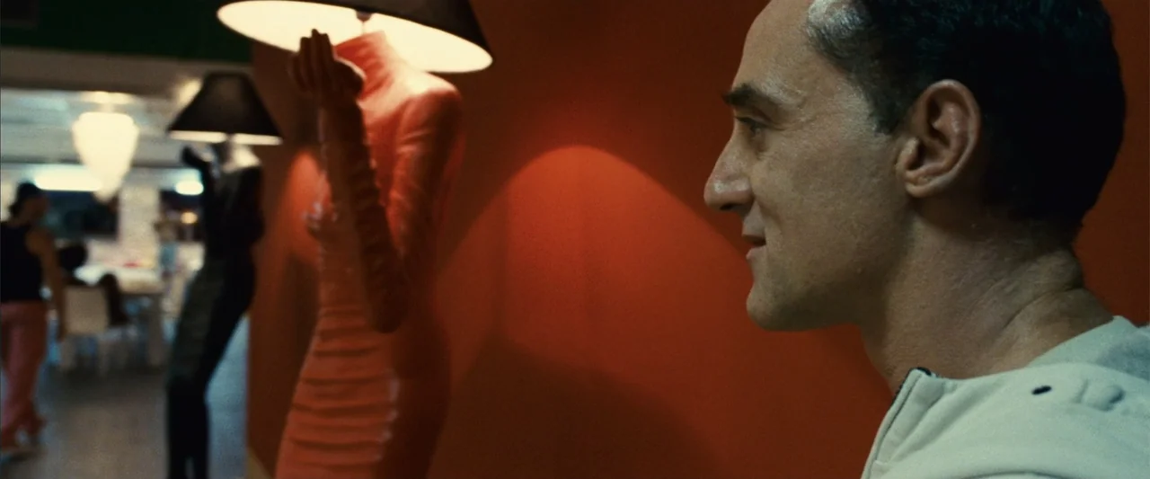 Reality (2023) movie still: medium — A man stands in a store with orange walls next to mannequin floor lamps. He looks to his right while positioned…