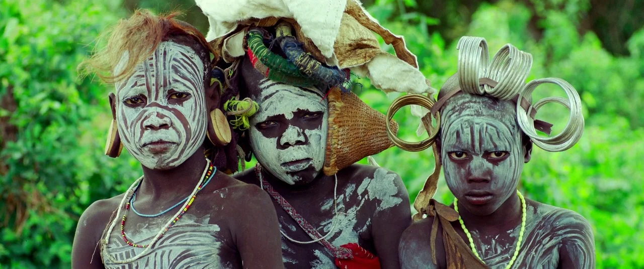 Samsara (2011) movie still: medium — Three children stand side-by-side in a lush green outdoor environment. They wear traditional white clay paint,…