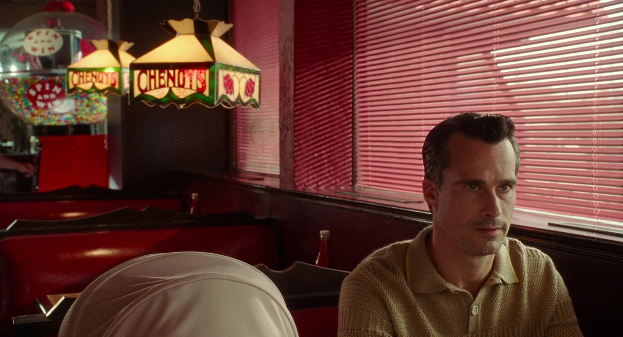 Babysitter (2022) movie still: medium — A diner booth features red vinyl seating and a Tiffany-style stained glass pendant lamp. A man sits at the…