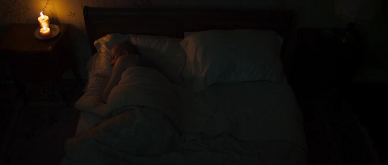 A Dark Song (2016) movie still: medium — A dark bedroom features a wooden bed frame with light-colored bedding and floral wallpaper. A man lies…