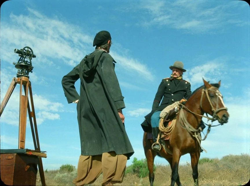 Jauja (2014) movie still: fullbody — An open desert landscape features a surveyor and a mounted cavalry soldier near surveying equipment. The bright…