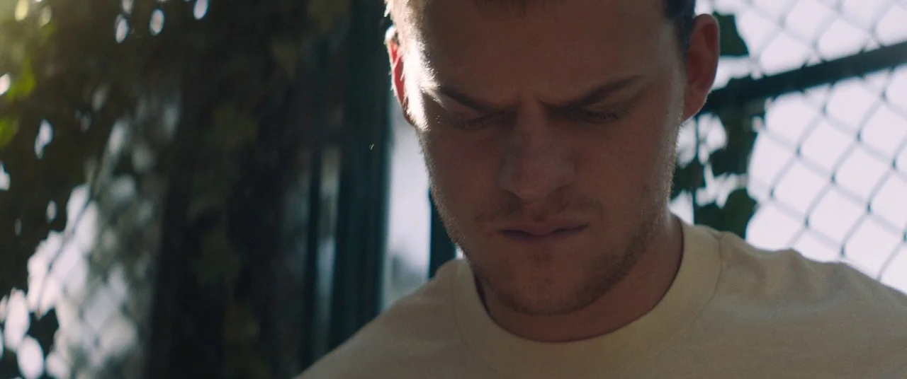 Honey Boy (2019) movie still: closeup — A close-up shot of a man standing before a chain-link fence overgrown with vines. He keeps his eyes closed…