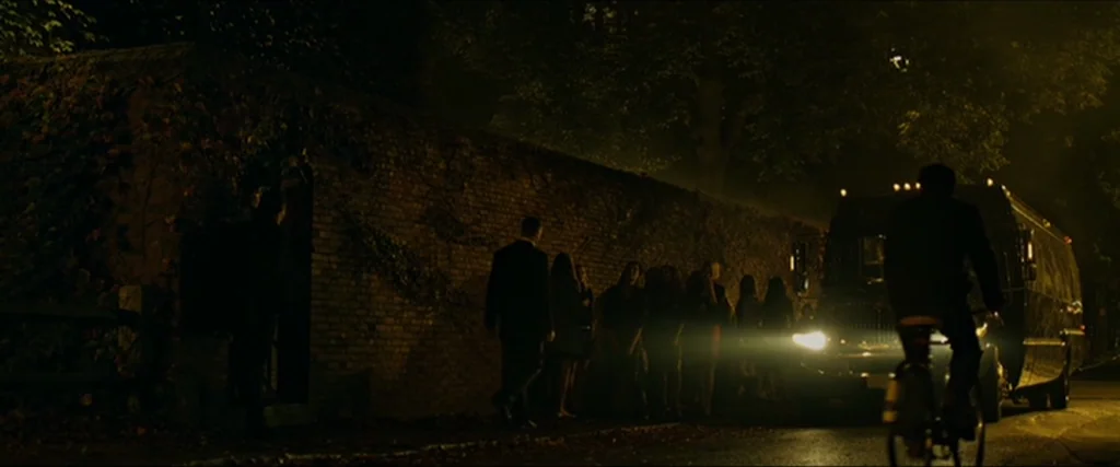 The Social Network movie still: wide — A long brick wall covered in vines lines a dark asphalt road at night. A group of people stands by the wall…