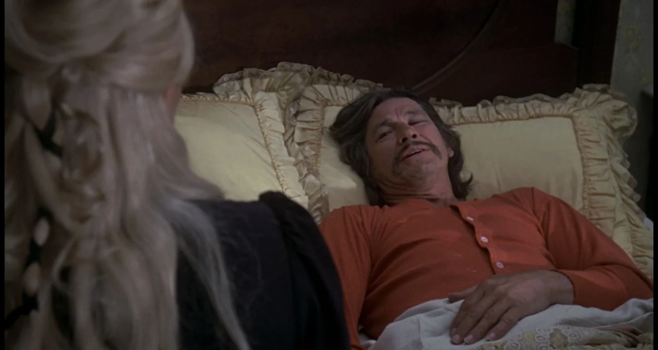 The White Buffalo (1977) movie still: medium — A bedroom contains a person in bed with yellow ruffled pillows and dark headboard. A woman sits on…