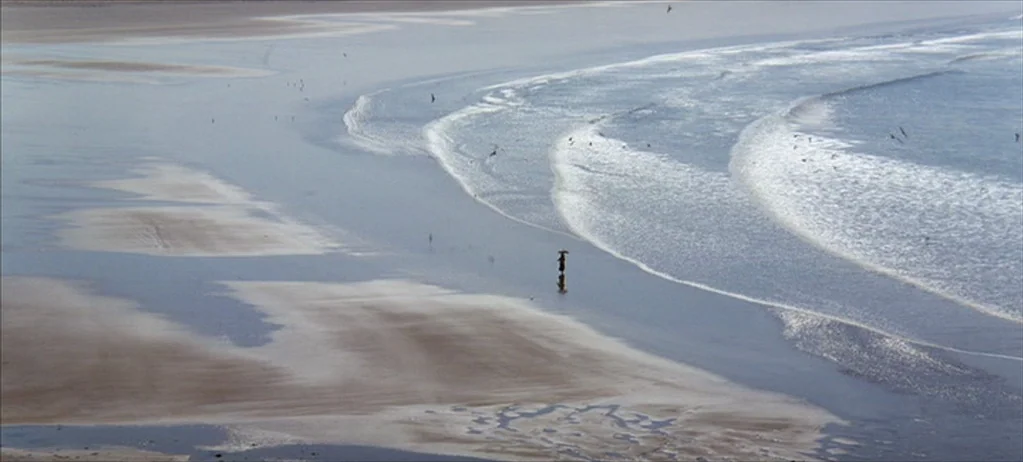 Ryan’s Daughter (1970) movie still: wide — An expansive, wet sandy beach stretches toward the rhythmic waves of a calm ocean. A lone figure walks…