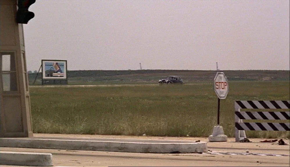 The Godfather (1972) movie still: wide — An open, flat landscape features a roadside traffic stop with a sign and barrier. A dark vintage car drives…