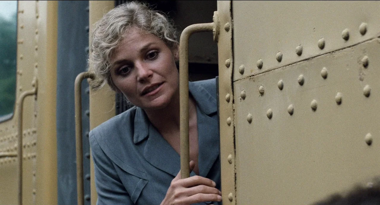 Desert Hearts (1985) movie still: closeup — A woman leans from the side of a yellow rivet-studded train car. The metallic exterior of the carriage…