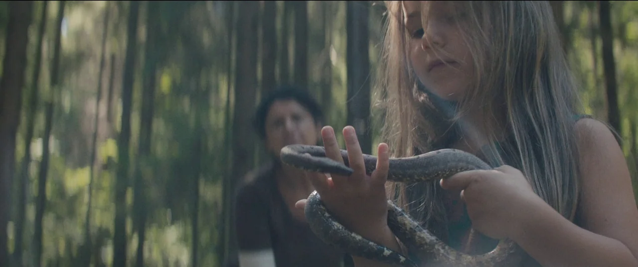 Nightsiren (2022) movie still: medium — A young girl holds a snake in a dense wooded area. A blurred woman stands in the background behind the girl.