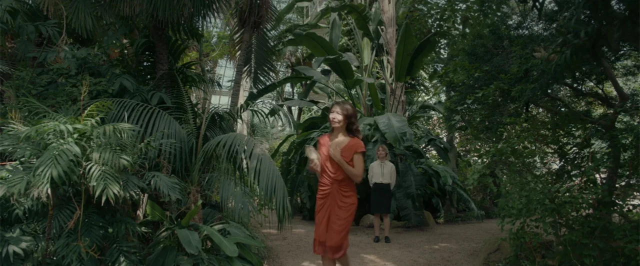 Peacock (2010) movie still: fullbody — A glass conservatory contains large tropical plants with a dirt path winding through foliage. A woman walks…