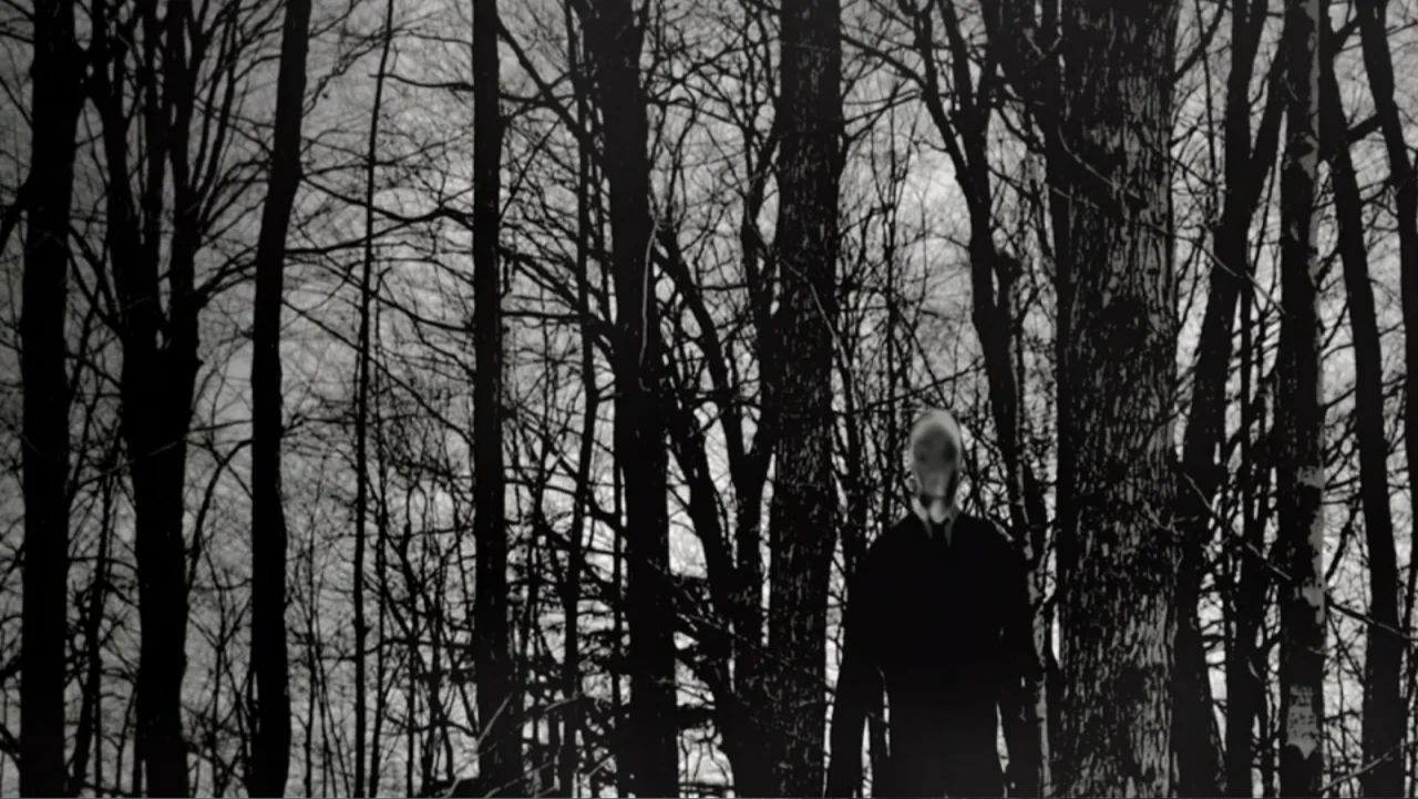 Beware the Slenderman (2016) movie still: medium — A dense grove of bare trees fills the frame with vertical branches and trunks. A tall figure in a…