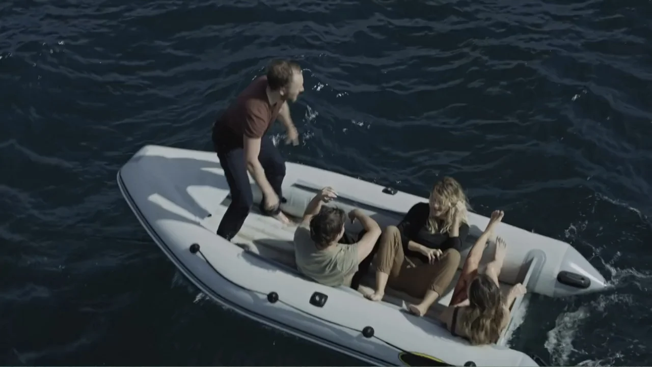 Capsized: Blood in the Water (2019) movie still: wide — An inflatable rubber boat floats on the deep blue surface of open water. Four people occupy…