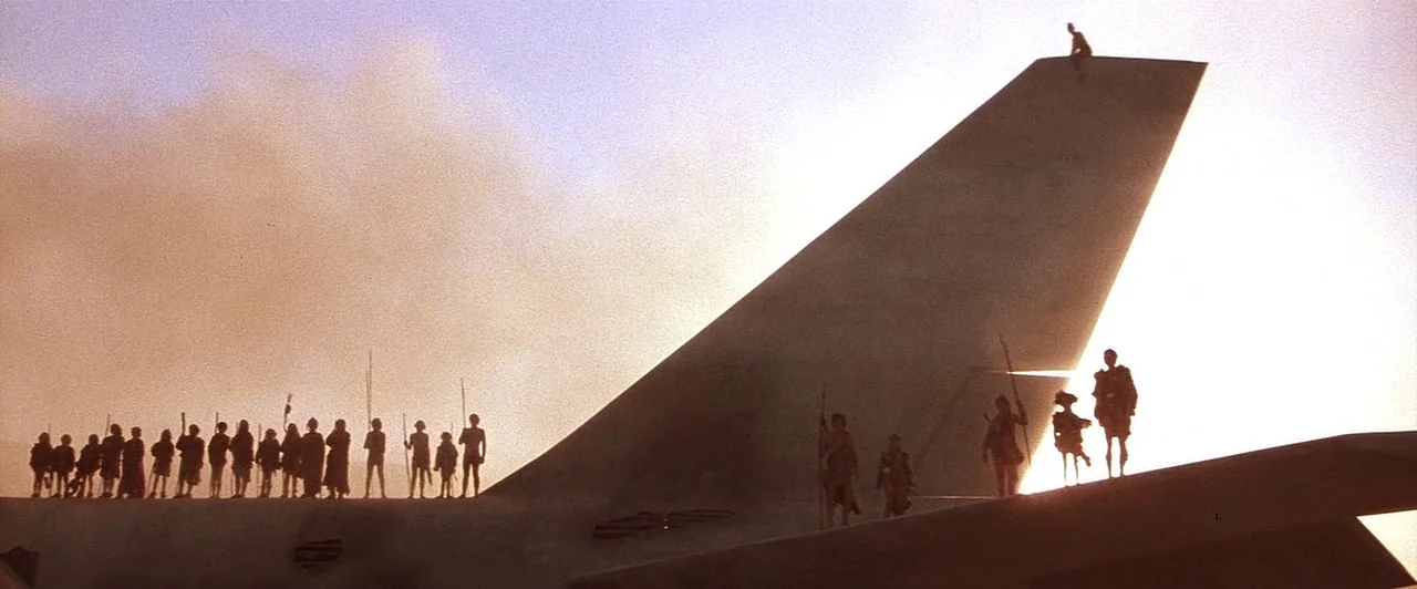 Mad Max Beyond Thunderdome (1985) movie still: wide — A group of people stands atop the wing of a grounded aircraft at sunset. The figures are…