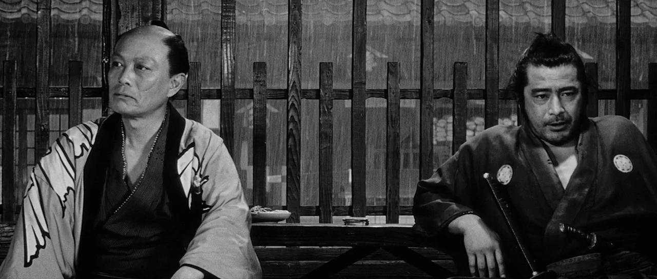 Yojimbo (1961) movie still: medium — A wooden veranda with vertical slats overlooks a rainy courtyard during the day. Two men in traditional…