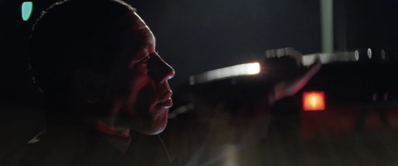 Colt 45 (2014) movie still: closeup — A man in darkness stands beside a road at night. Red light illuminates the side of his face as he looks forward.