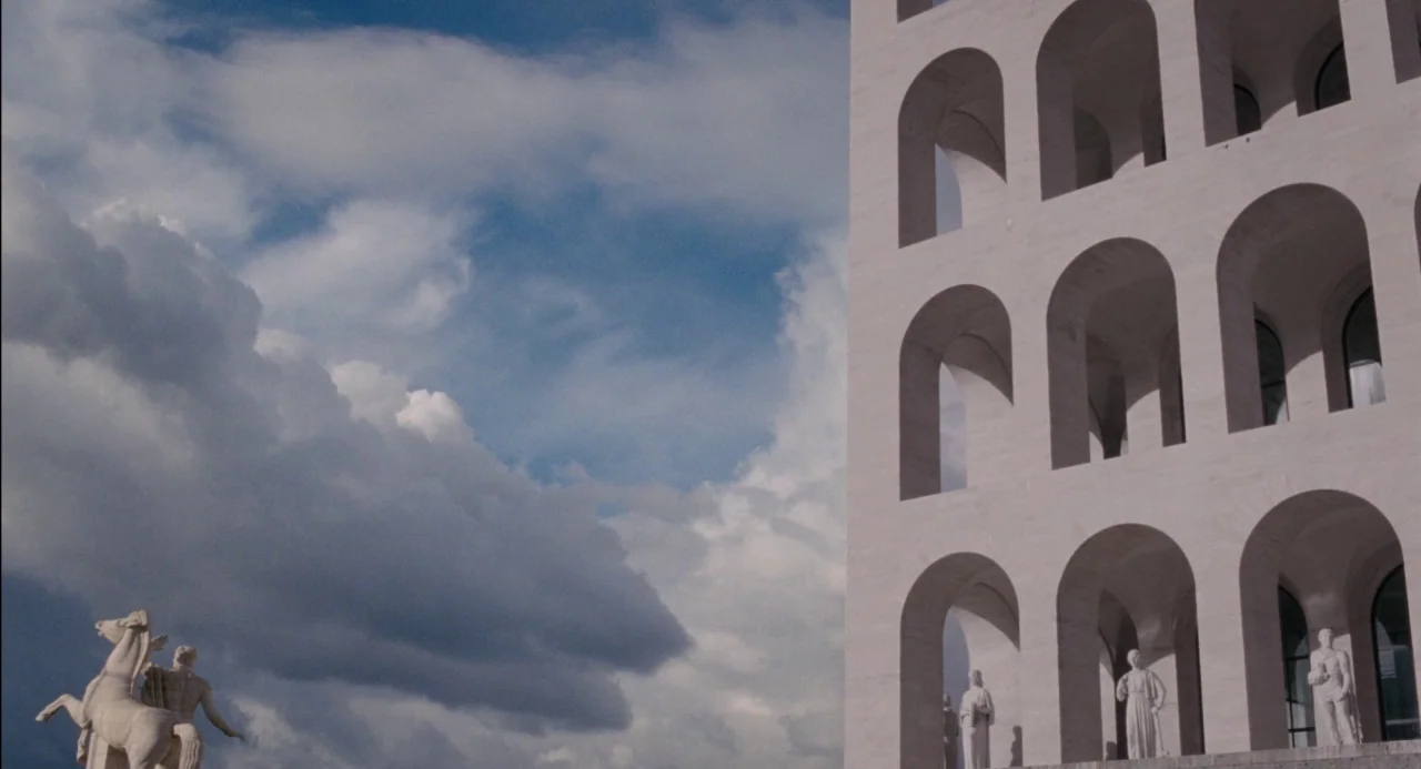 Pasolini (2014) movie still: establishing — A massive, multi-story travertine building with stacked rows of arches dominates the frame. Statues…