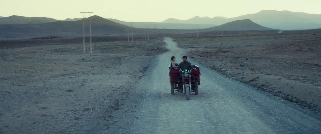 Animalia (2023) movie still: wide — A desolate desert landscape with rolling hills and a straight gravel path. A man and a woman sit on a red…