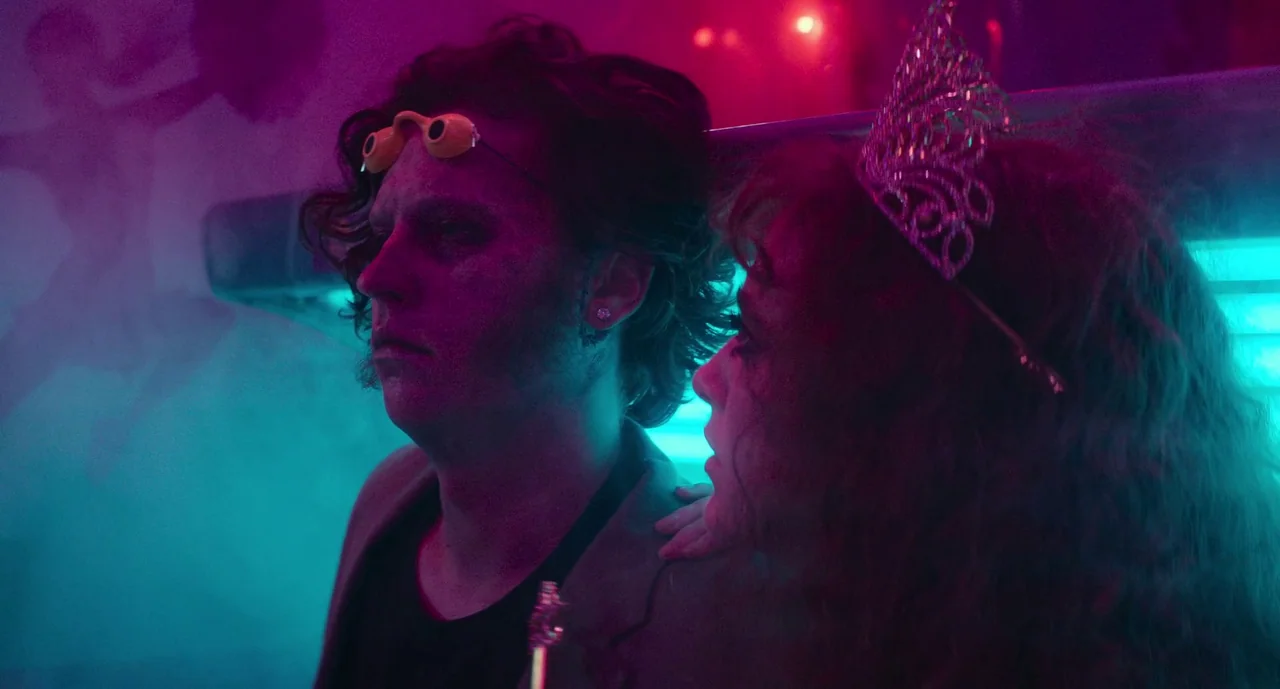 Lisa Frankenstein (2024) movie still: medium — A crowded venue interior with heavy fog and intense dual-colored neon lighting. Two people stand in…