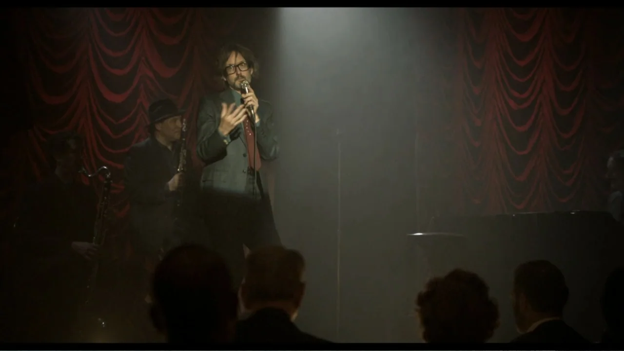 Six by Sondheim (2013) movie still: medium — A performance space features a dark stage backdrop with deep red gathered curtains. A man in a suit…