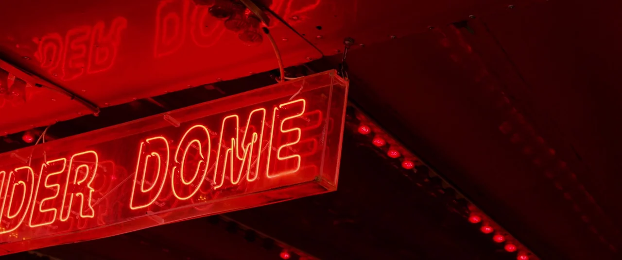 Bull (2021) movie still: closeup — A close-up view of a rectangular red neon sign spelling the word DOME. The sign hangs against a dark background,…