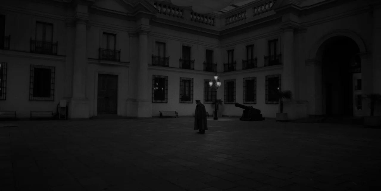 El Conde (2023) movie still: wide — A dark, expansive stone courtyard surrounds a large neoclassical government building at night. A lone uniformed…