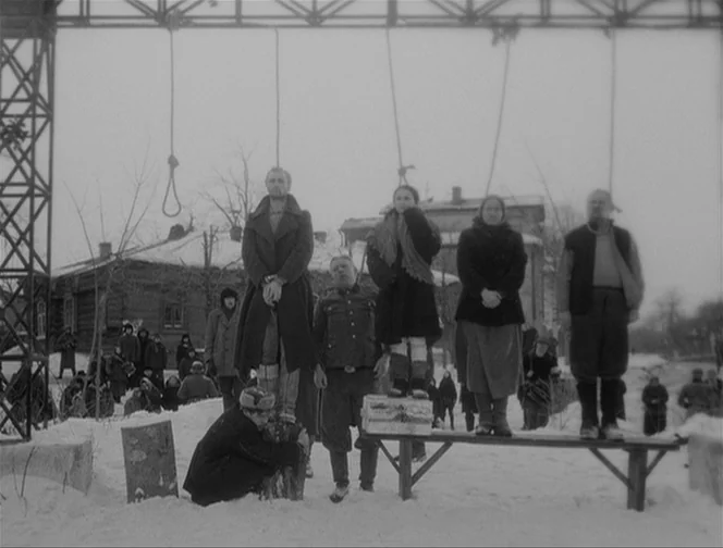 The Ascent (1977) movie still: fullbody — A snow-covered village square features a large metal structure with multiple nooses hanging down. Several…