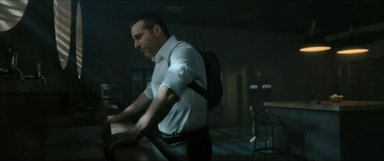 Kraven the Hunter (2024) movie still: medium — A dimly lit interior room with a dark counter and circular wall cutouts. A man in a white shirt…