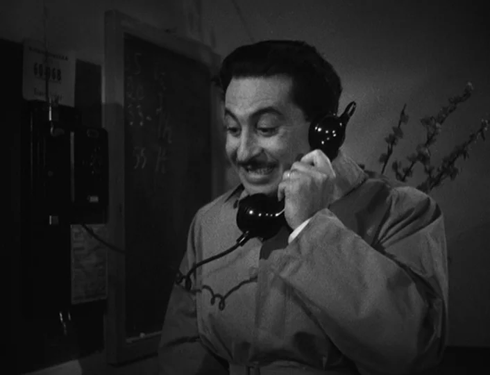 The White Sheik (1952) movie still: medium — A man stands in an office with a blackboard covered in chalk numbers. He holds a vintage wall-mounted…