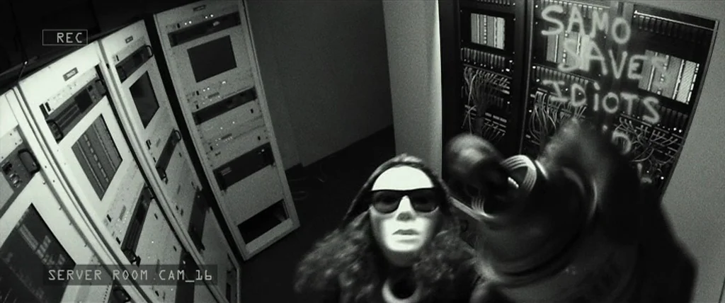 Dope (2015) movie still: medium — A server room contains several tall black racks with blinking electronic components and visible cables. A masked…