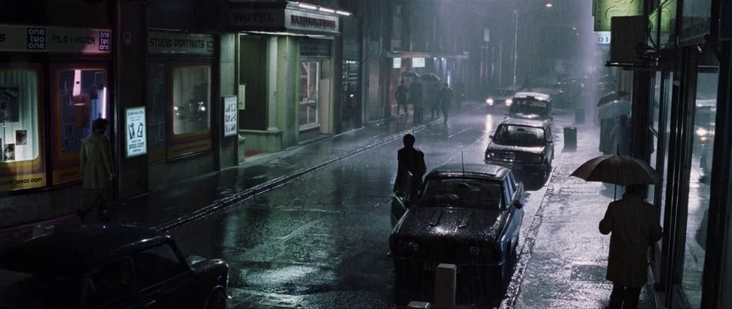 Munich (2005) movie still: wide — A rain-drenched city street features parked cars and storefronts with vintage 1970s signage. Pedestrians walk…