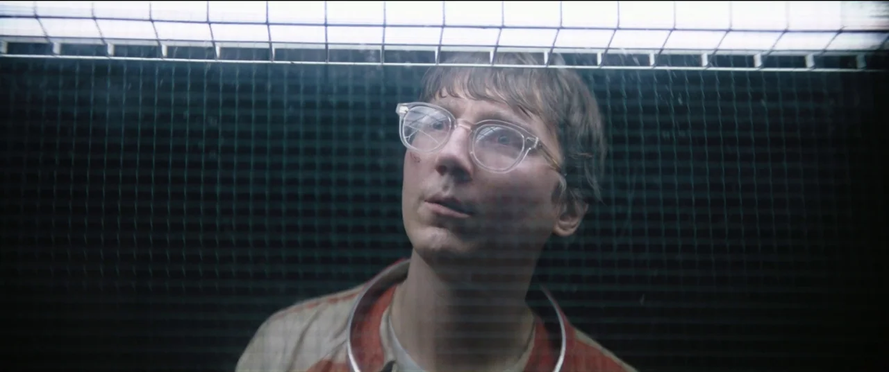The Batman (2022) movie still: closeup — A stark room features a heavy wire security screen separating two sides. A man stares upward with a neutral…