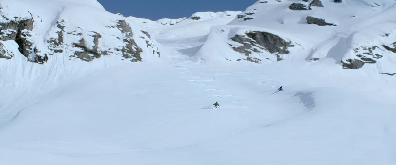 Force Majeure (2014) movie still: wide — A vast, steep snow-covered basin is flanked by rugged, rocky cliffs under blue skies. Two skiers descend…