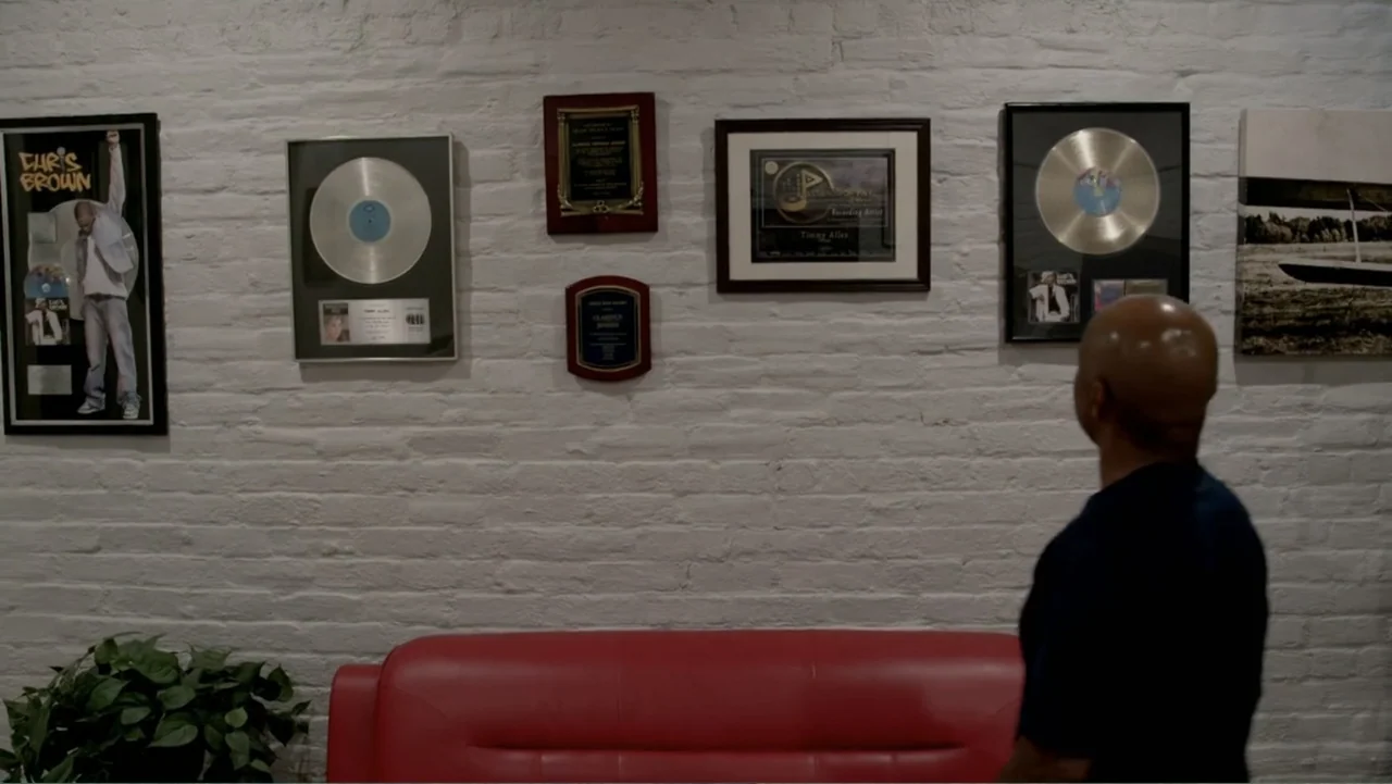 Chris Brown: A History of Violence (2024) movie still: medium — A white brick wall is decorated with several framed music industry awards and…